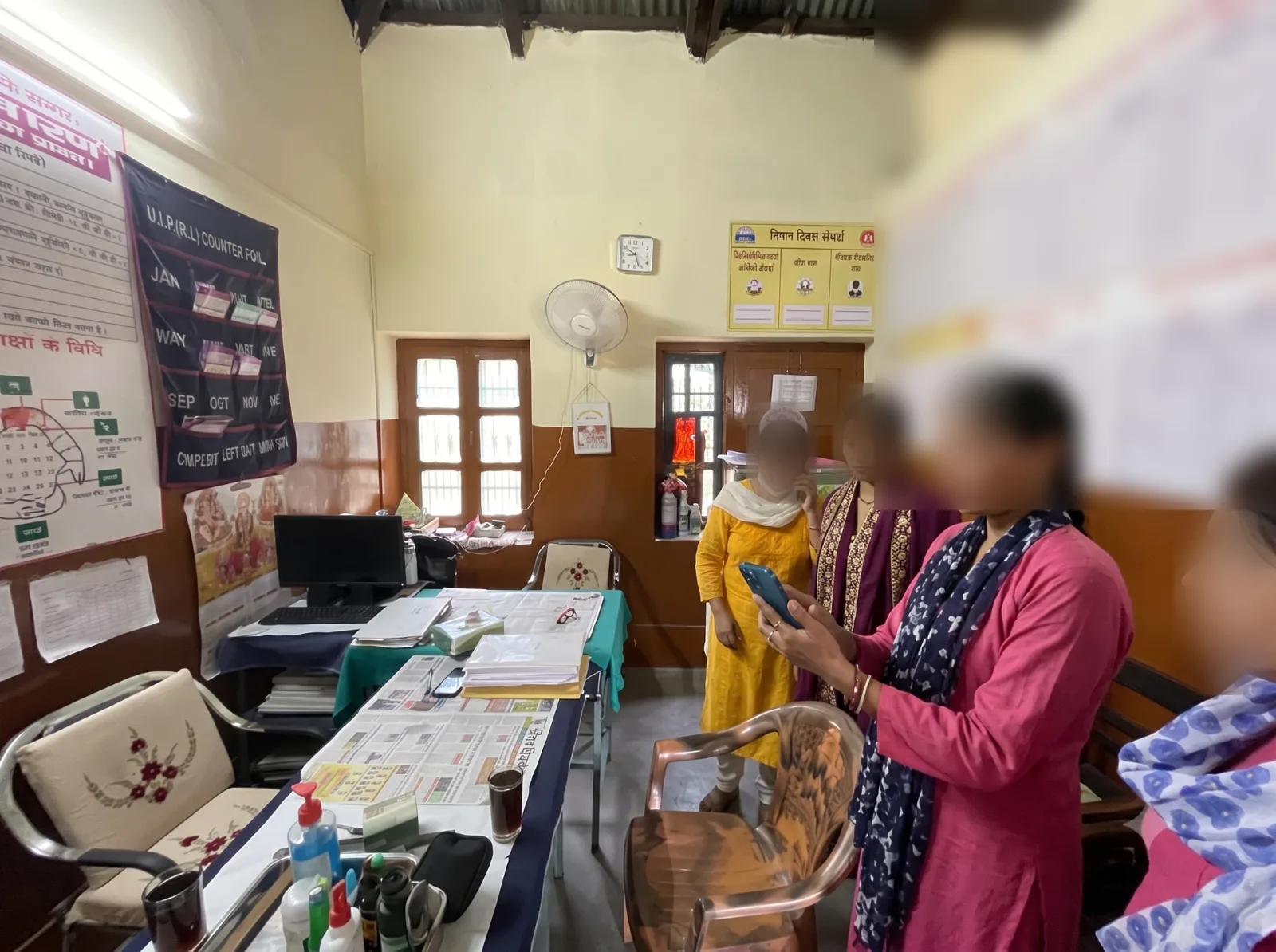 Four health workers in a sub-centre room, one holding up a phone to try the prototype while the others look on, faces blurred